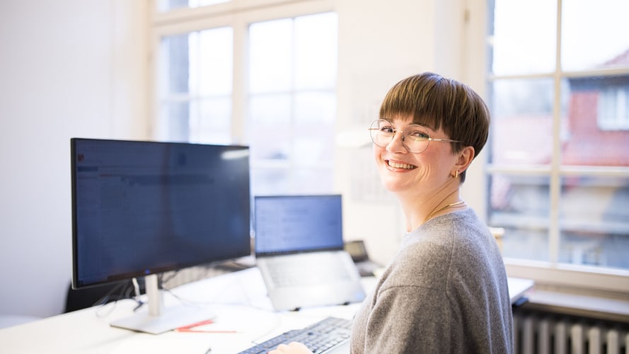 Eine lächelnde Frau mit Brille und kurzem Haar sitzt an einem Schreibtisch mit zwei Bildschirmen und einem Laptop. Sie blickt über die Schulter in die Kamera. Der helle Büroraum hat große Fenster mit Blick auf ein Gebäude mit roten Dachziegeln.